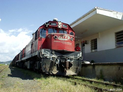 Locomotiva vermelha da América Latina Logística parada junto ao prédio da estação de Lages, com a composição atrás.