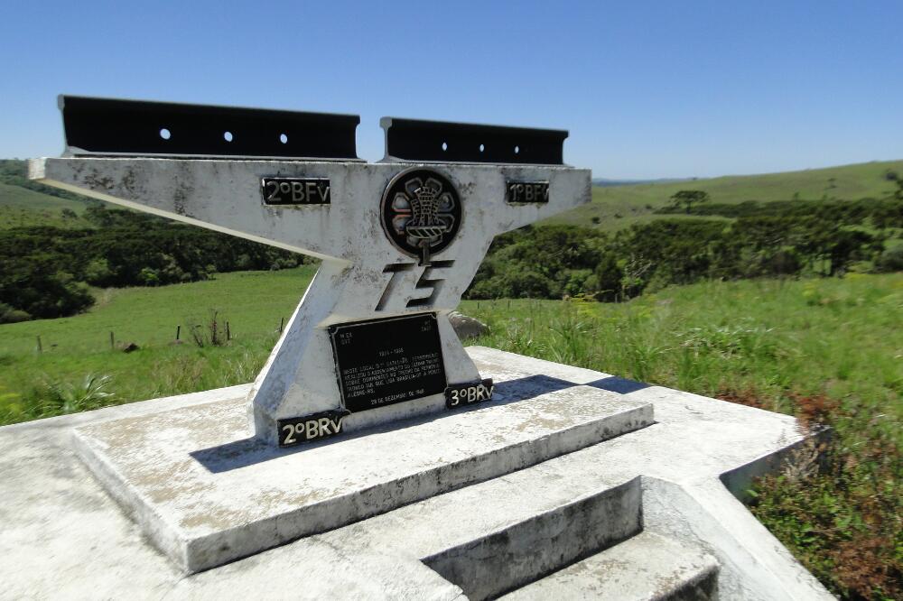 Monumento de concreto com duas barras de trilho fixadas no topo e as insígnias dos batalhões ferroviários, em campo aberto.