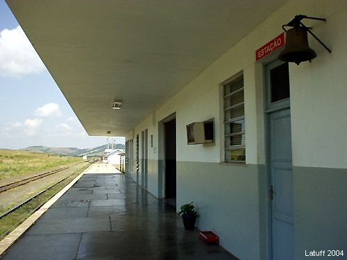 Plataforma vazia da estação de Lages sob a marquise, com o sino e a placa ESTAÇÃO na parede, a linha férrea e a serra ao fundo.